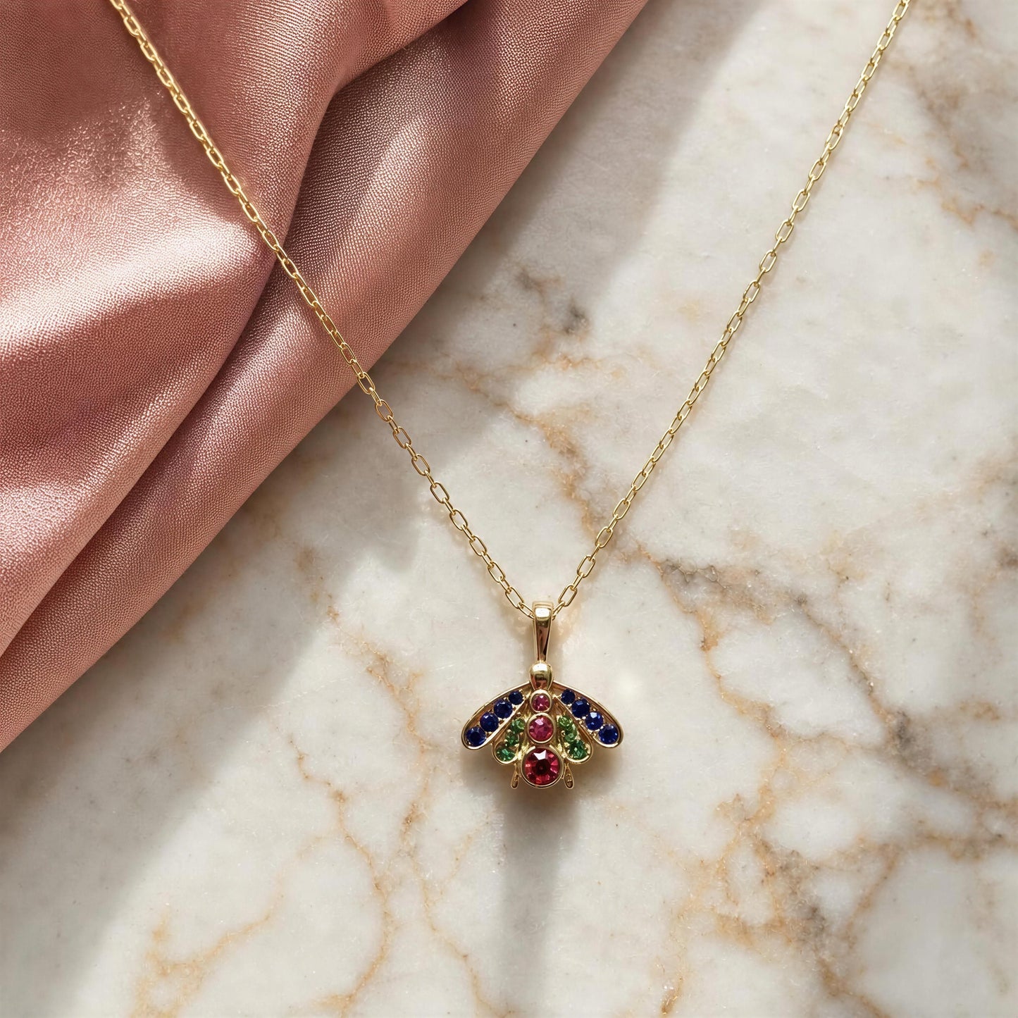 A gold chain necklace with a butterfly pendant, decorated with colorful enamel wings in blue, green, and red, displayed on a white marble surface next to a pink satin fabric.