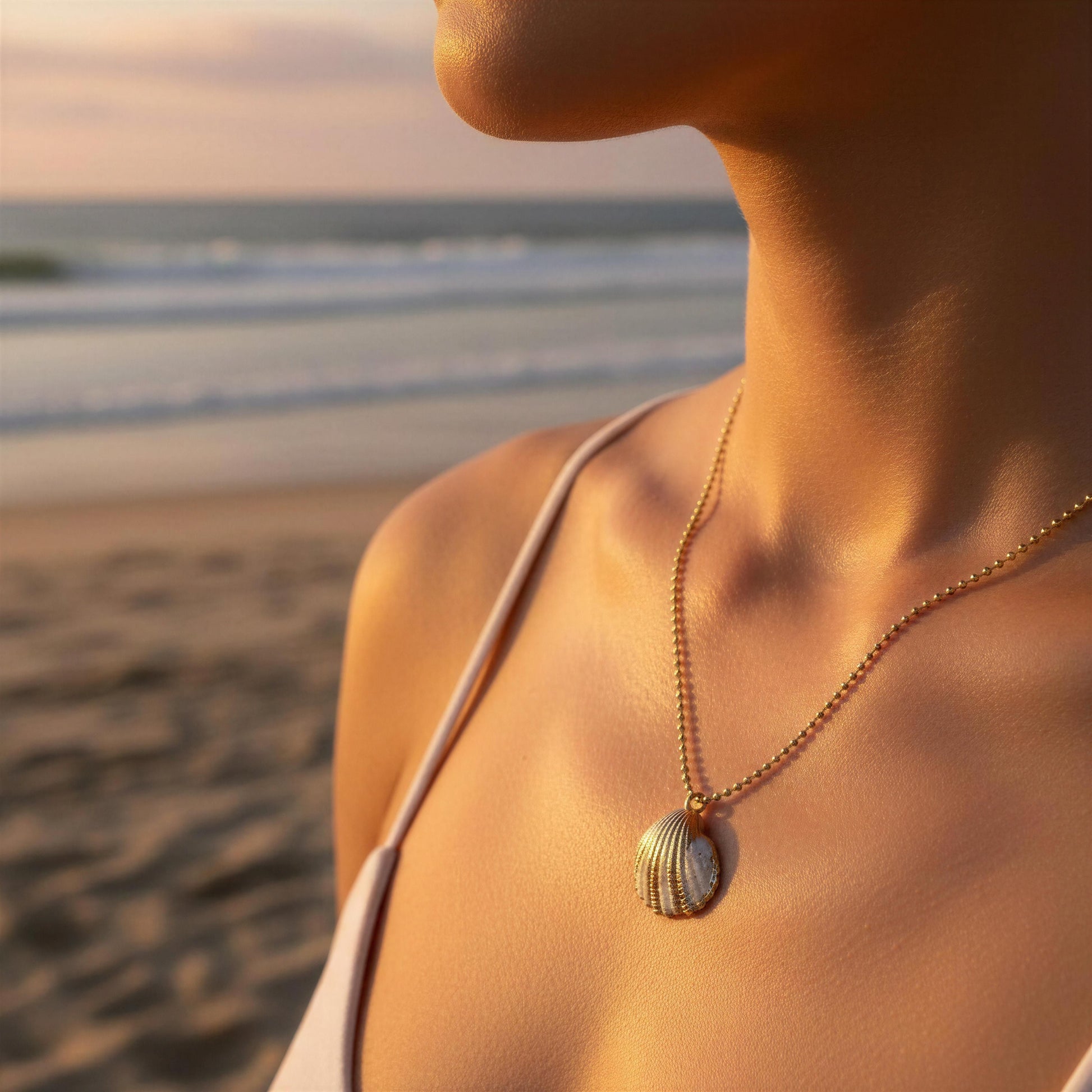 Woman wearing a gold necklace with a shell pendant on a beach at sunset.