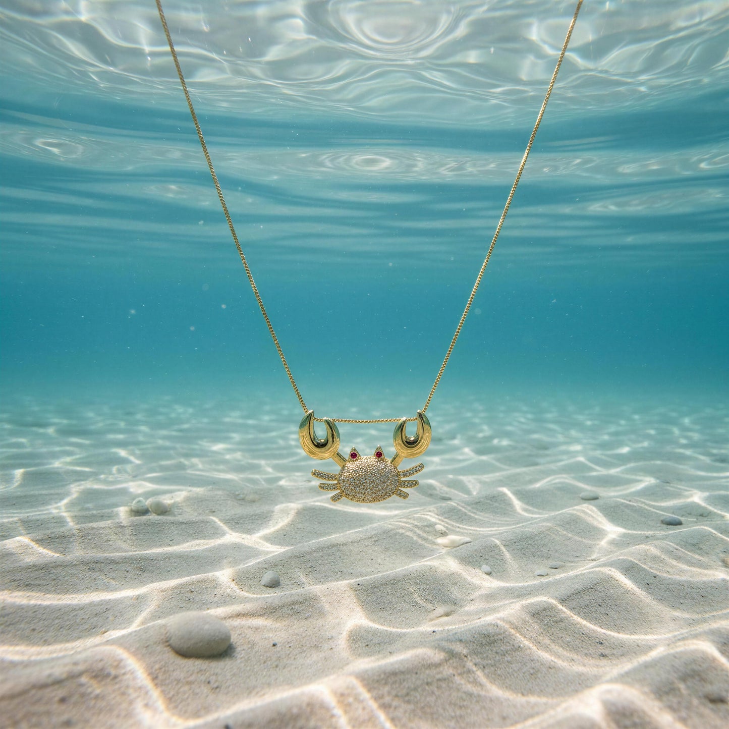 The Golden Tides Crab Necklace from Chica Chica Jewelry  underwater on a sandy ocean floor.