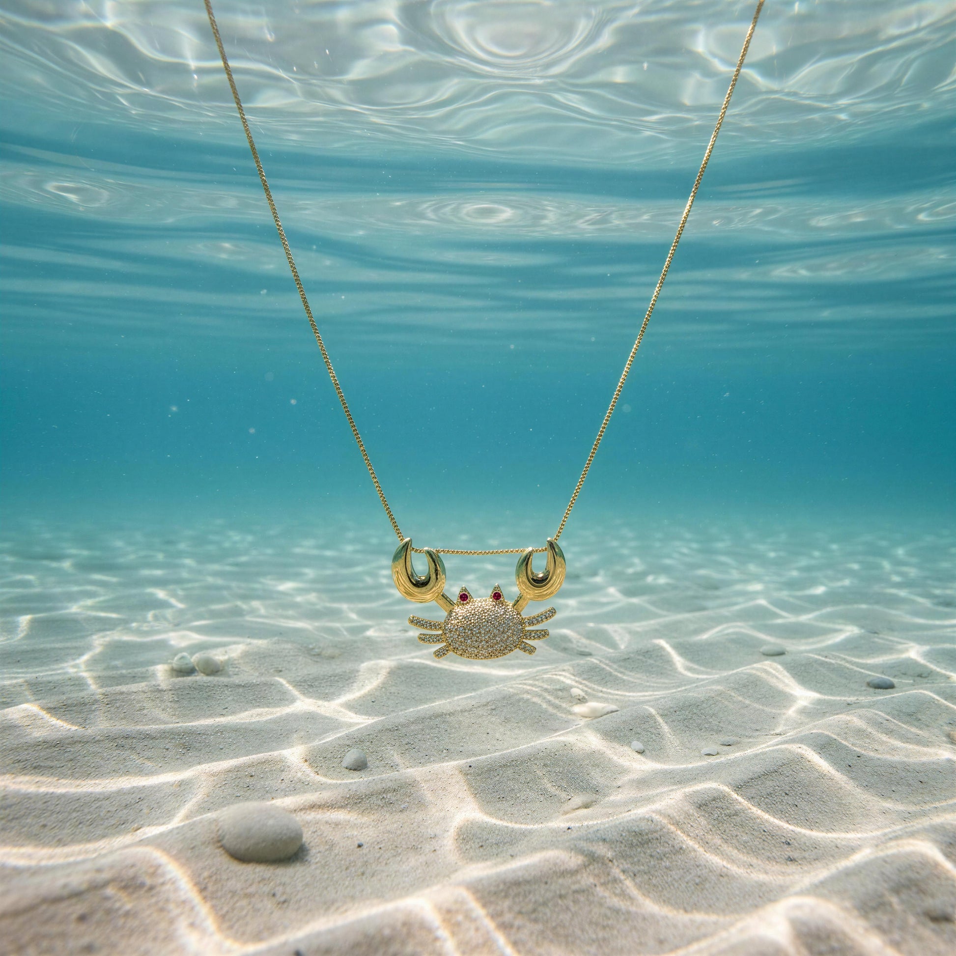 The Golden Tides Crab Necklace from Chica Chica Jewelry  underwater on a sandy ocean floor.