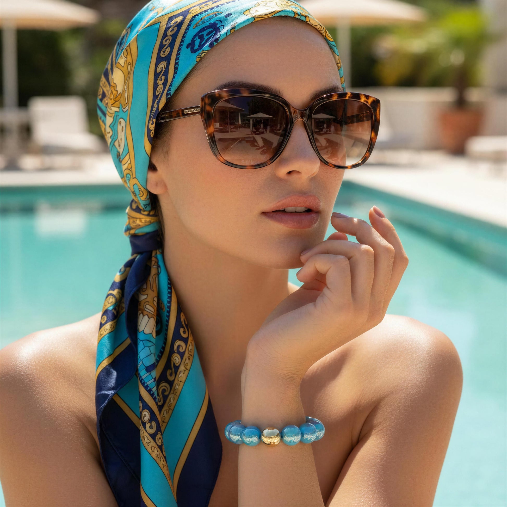 Woman wearing a colorful headscarf and sunglasses by a poolside and the Greek Glow Bracelet from Chica Chica Jewelry.