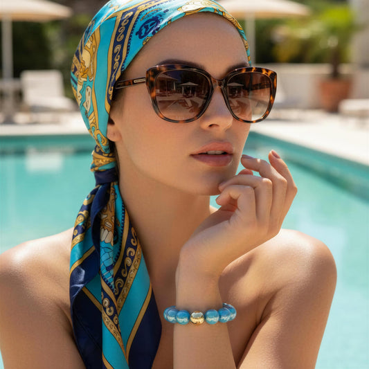 Woman wearing a colorful headscarf and sunglasses by a poolside and the Greek Glow Bracelet from Chica Chica Jewelry.