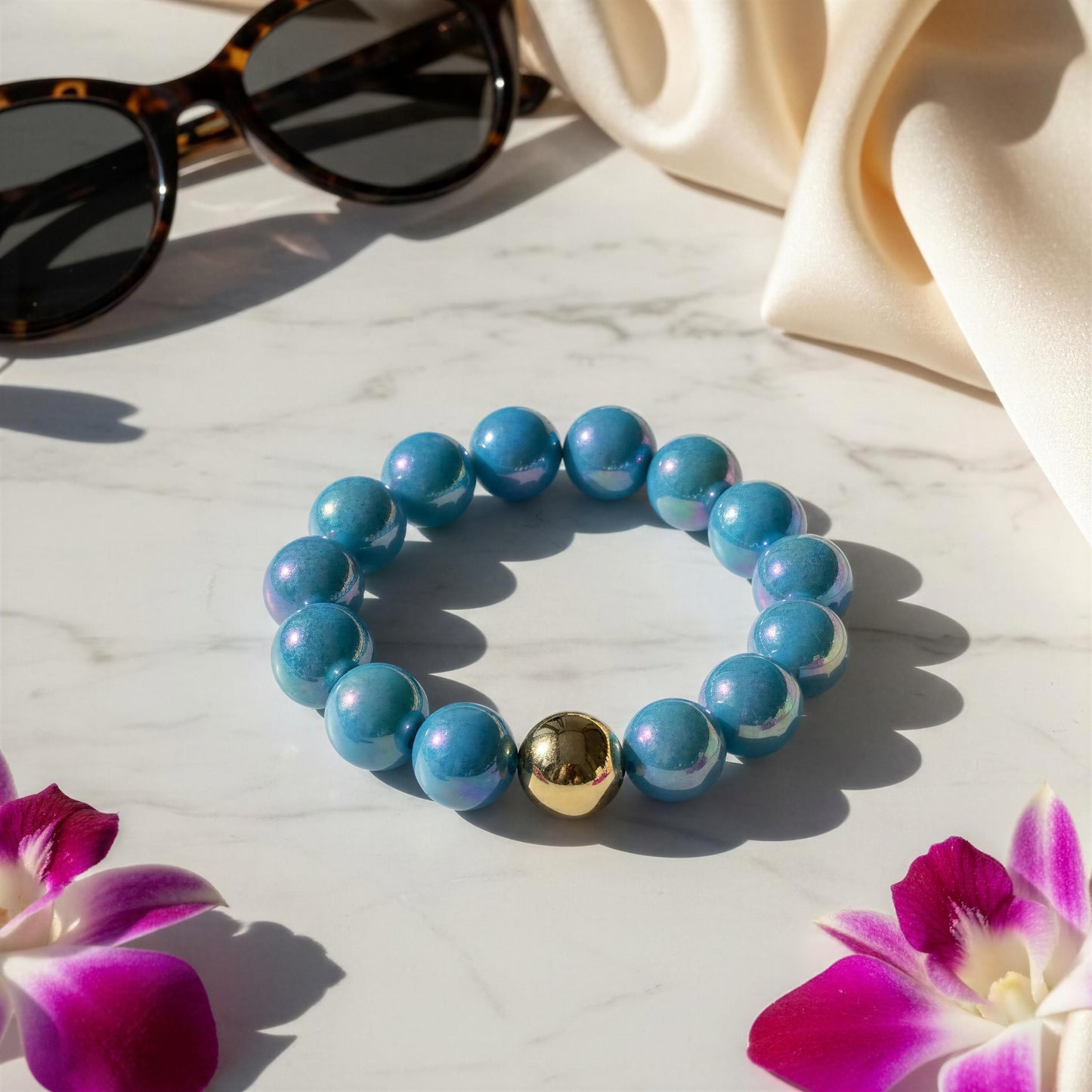 Blue beaded bracelet with a gold bead on a marble surface with sunglasses and flowers.
