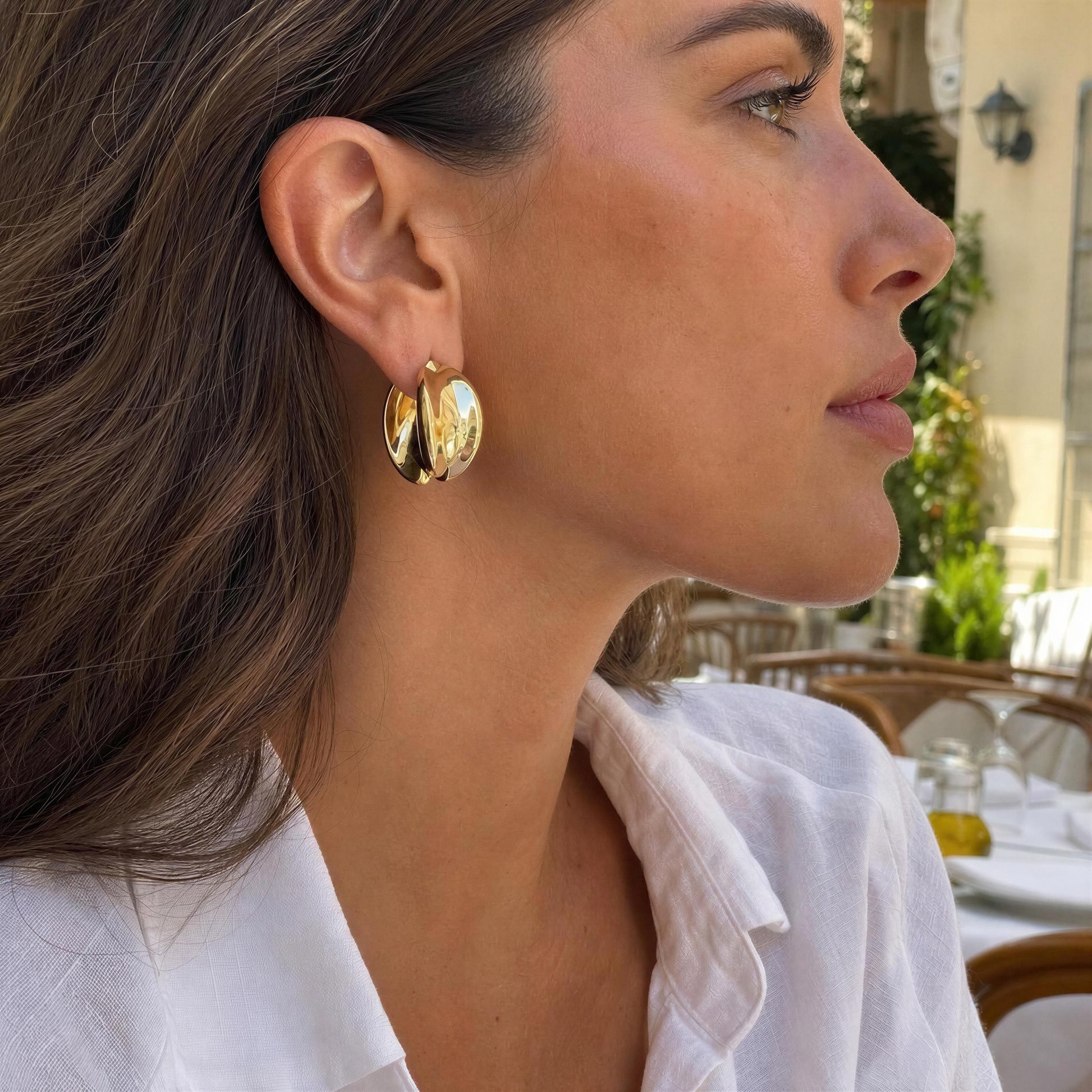 A woman in a white collared shirt wearing chunky, polished gold hoop earrings, photographed in a bright outdoor setting.