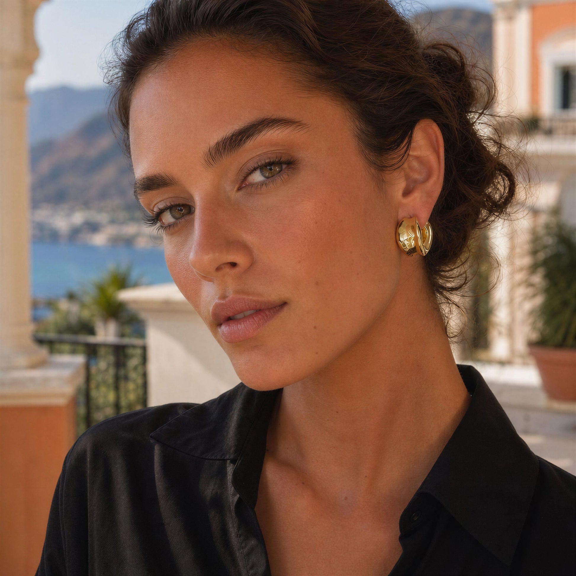 A close-up portrait of a woman wearing the Isadora gold hoop earring, from Chica Chica Jewelry, set against a blurred outdoor background of a coastal town and mountains.