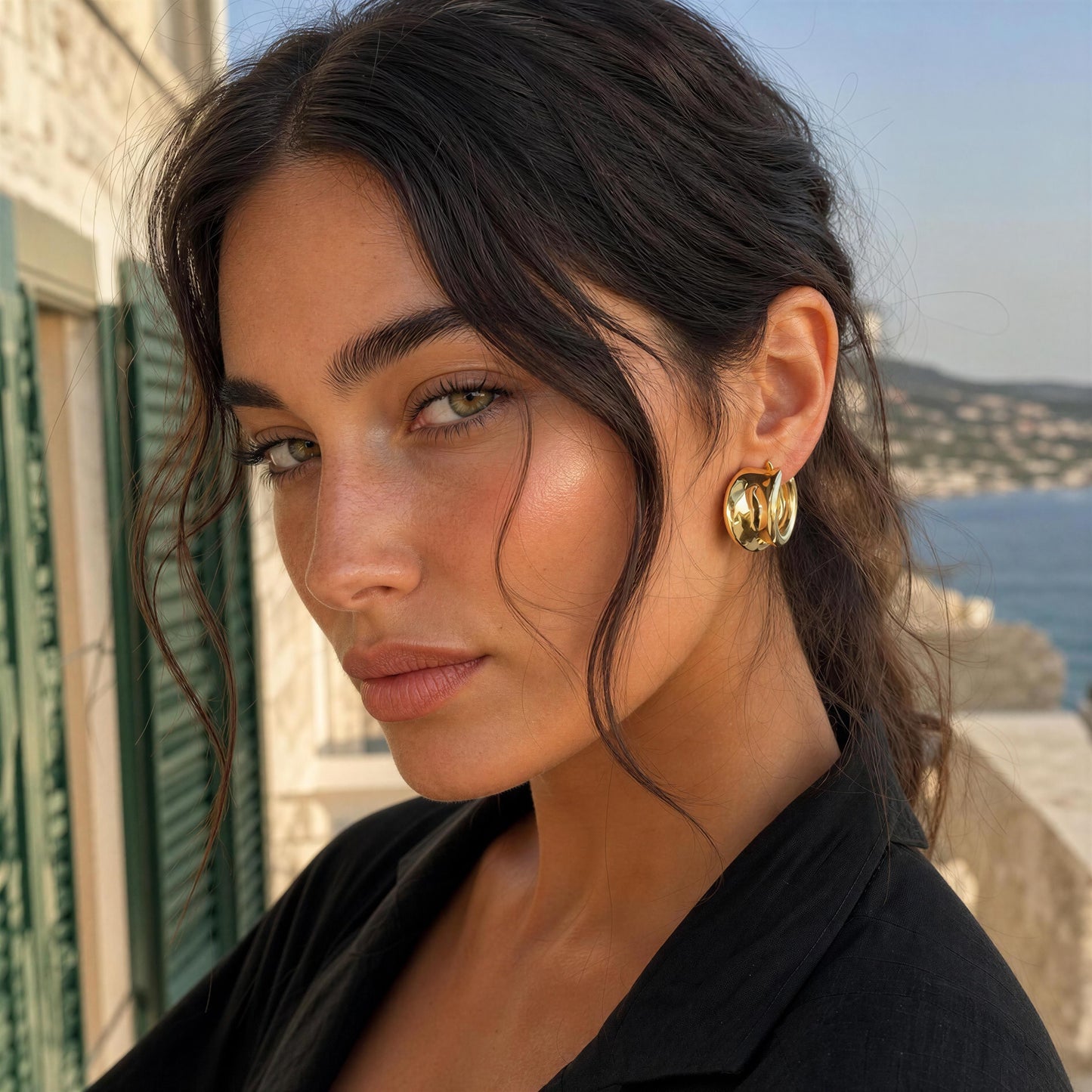 A close-up lifestyle shot of a woman with dark hair wearing the Isadora Hoops from Chica Chica Jewelry, a chunky gold hoop earring, set against a scenic coastal background with a stone wall and blue sea.