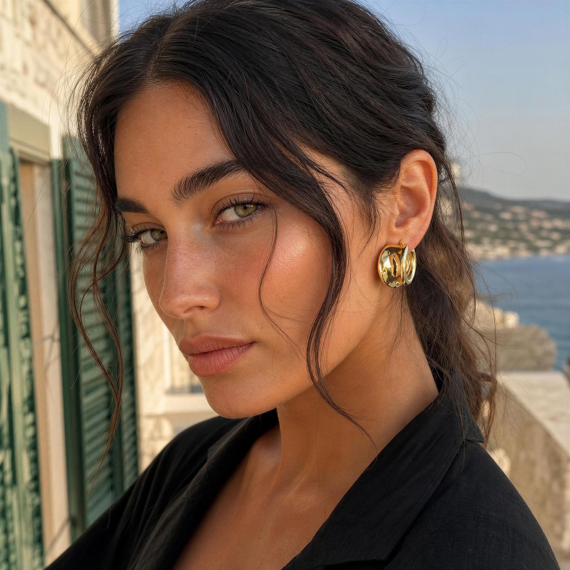 A close-up lifestyle shot of a woman with dark hair wearing the Isadora Hoops from Chica Chica Jewelry, a chunky gold hoop earring, set against a scenic coastal background with a stone wall and blue sea.