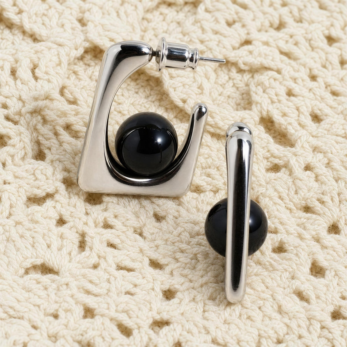 The Kara_Earrings from Chica Chica Jewelry; a pair of modern geometric hoop earrings with a polished silver-tone finish and a smooth black spherical accent, displayed on a textured cream crochet fabric background.