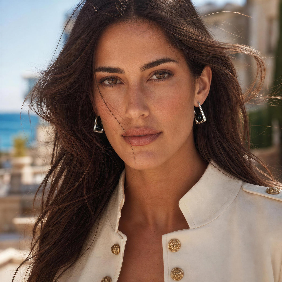 A close-up portrait of a woman wearing  the Kara Earrings from Chica Chica Jewelry, geometric silver earrings with black inlay, paired with a cream military-style jacket. The background is a softly blurred outdoor coastal setting.