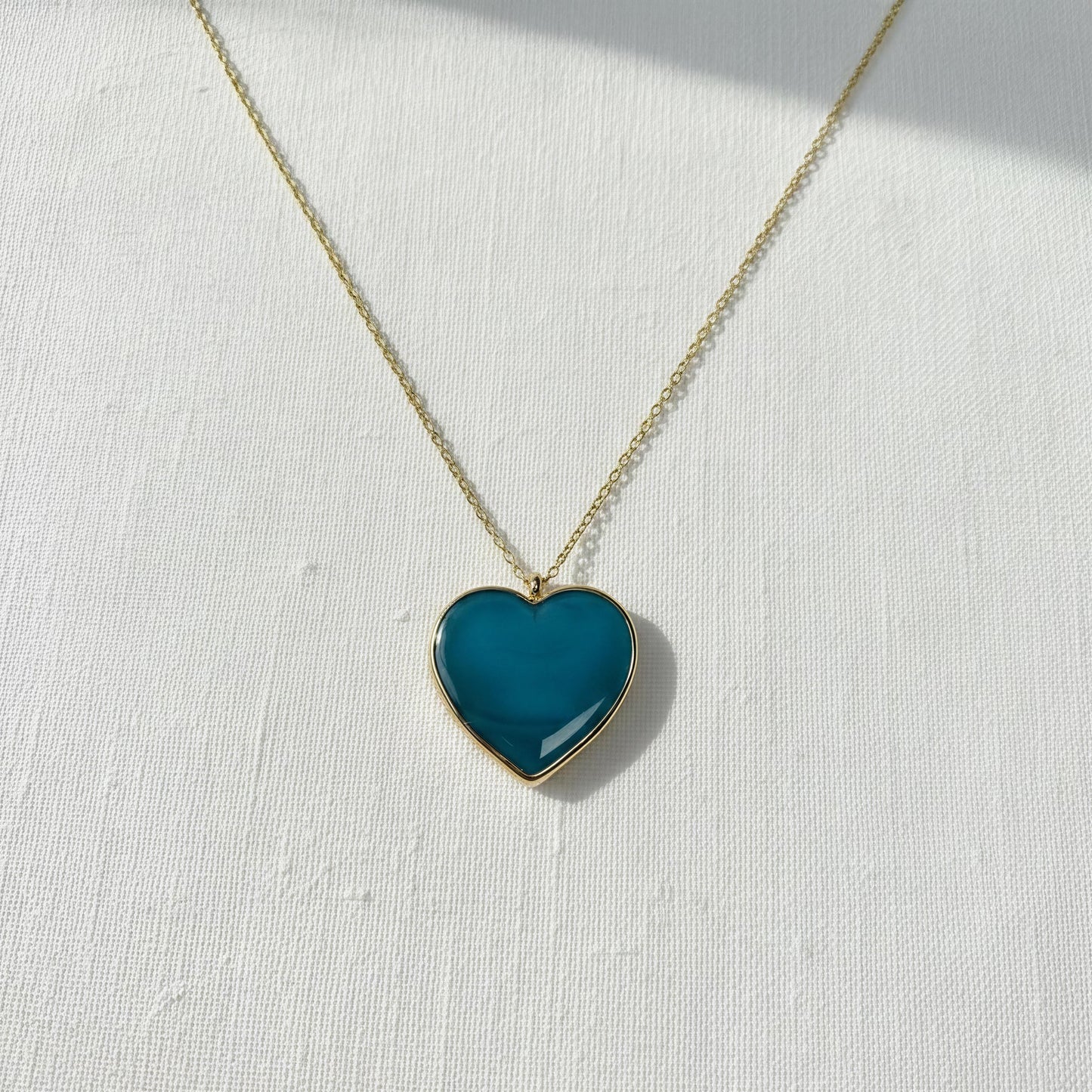Teal heart-shaped pendant on a gold chain against a white background