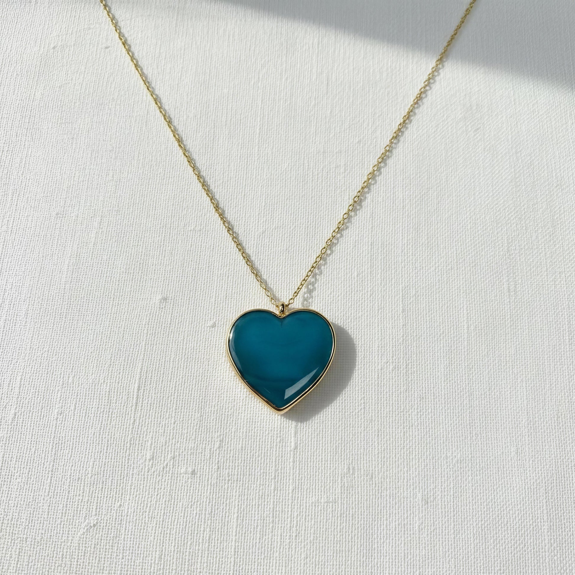 Teal heart-shaped pendant on a gold chain against a white background