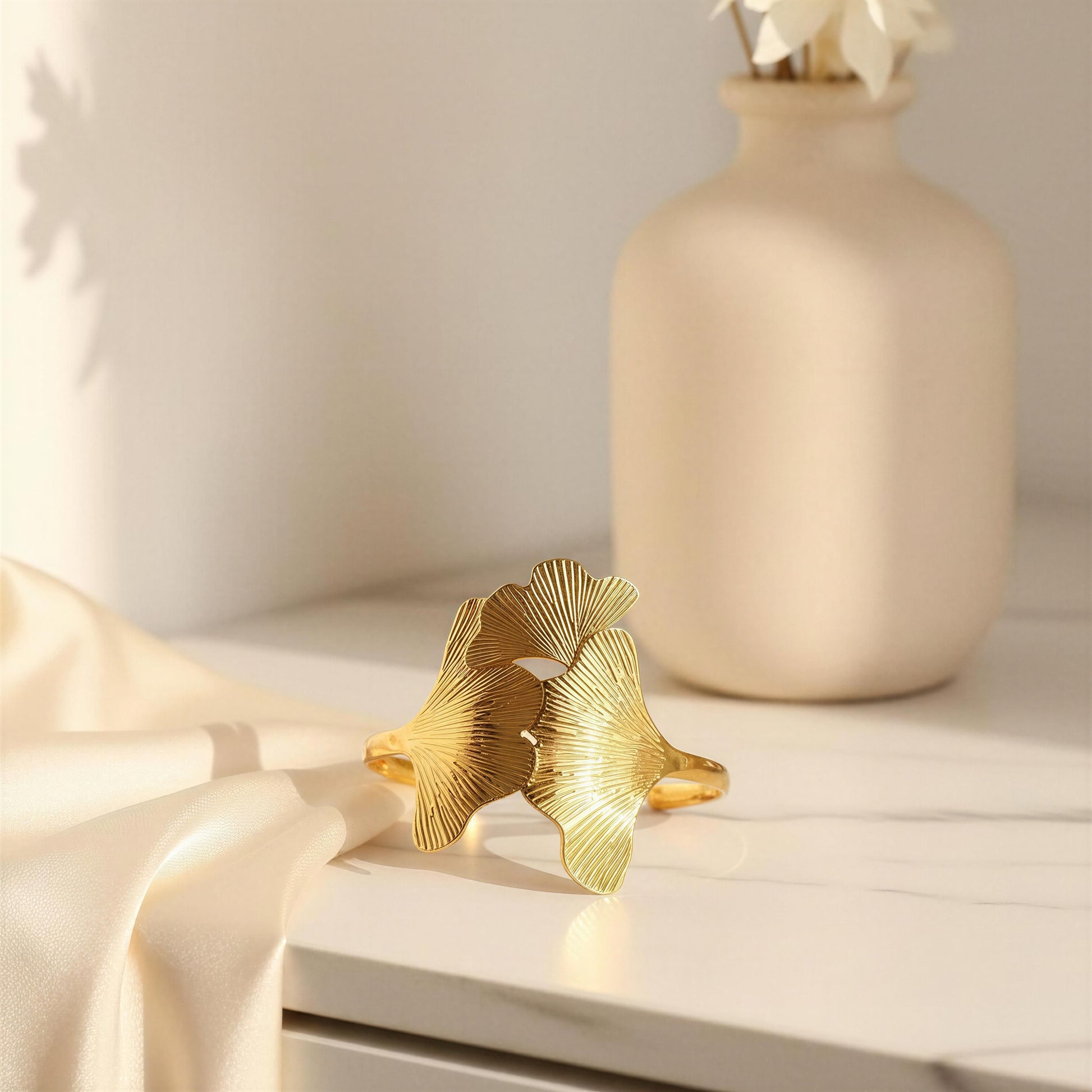 Gold leaf-shaped cuff bracelet from Chica Chica Jewelry on a white surface with a beige vase in the background. 