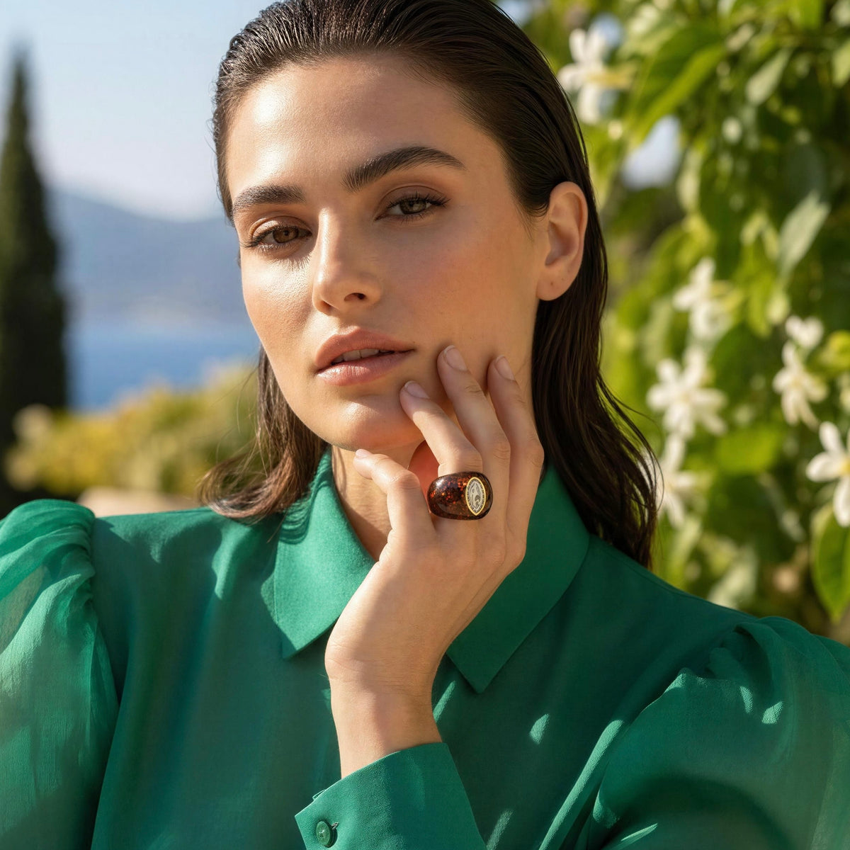 A woman in a green emerald organza blouse wearing the Lola_Ring_Toffee from Chica_Chica_Jewelry, posed outdoors with green foliage and a blurred scenic background.