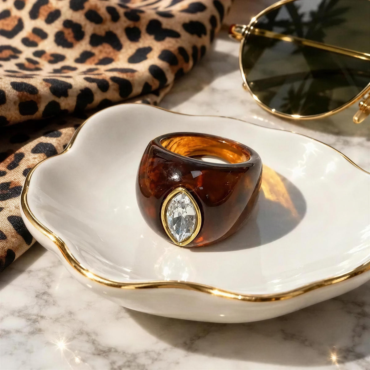 The Lola Ring in Toffee color set in a jewelry dish next to a silk animal print scarf and aviator sun
