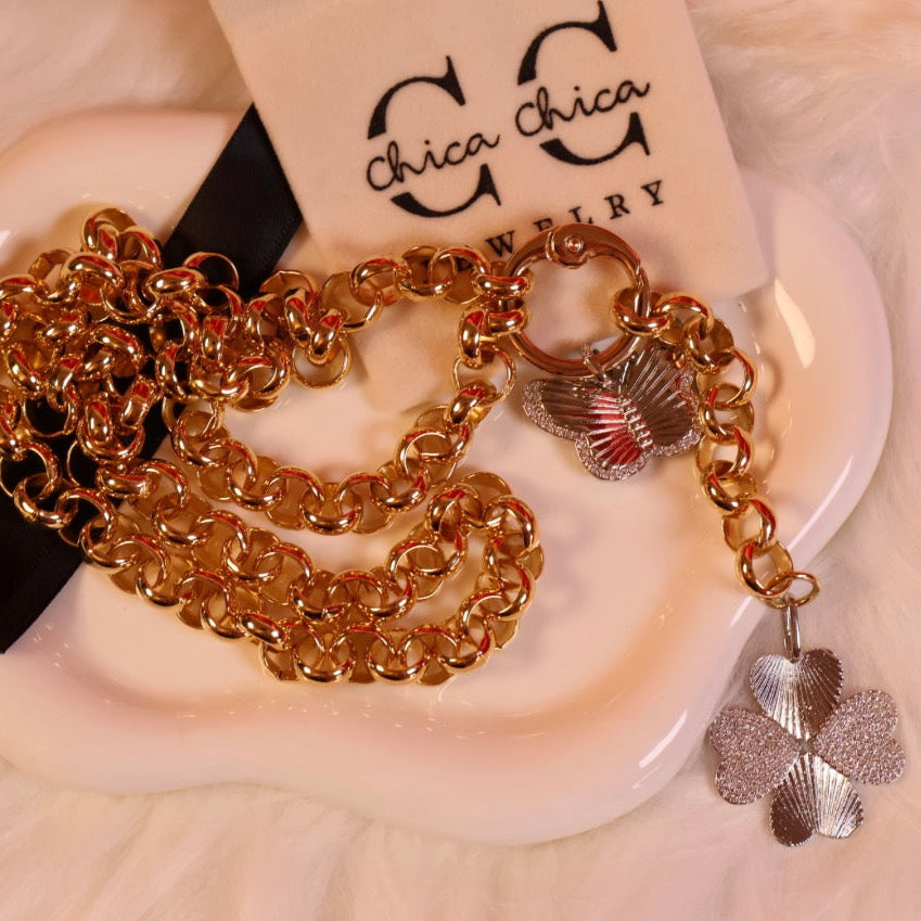Gold chain bracelet with charms on a white dish, branded 'Chica Chica Jewelry'.