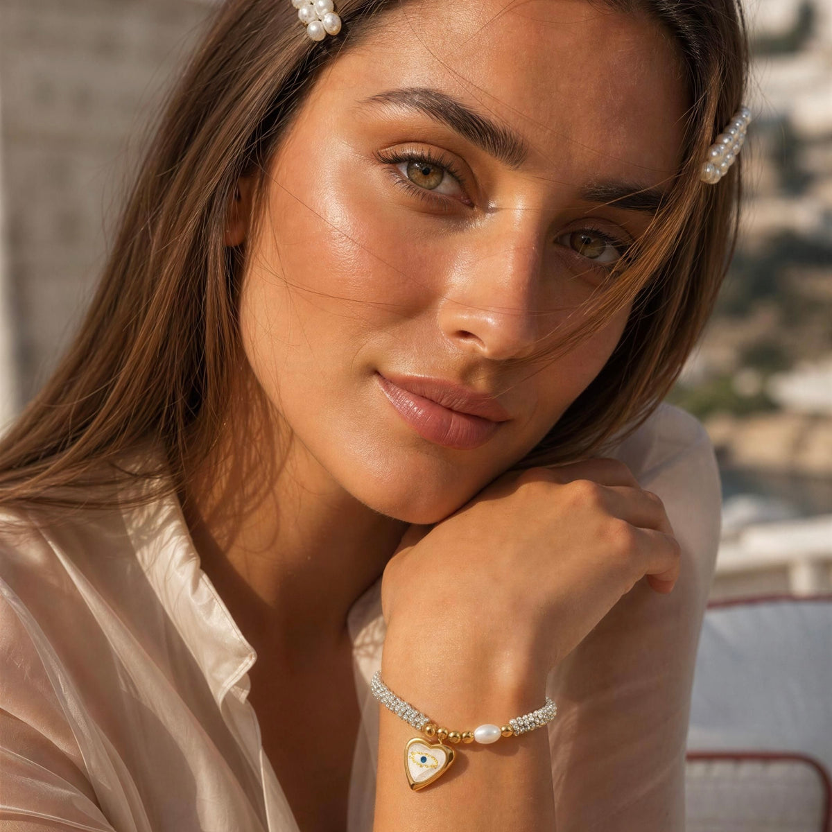A close-up lifestyle shot of a woman wearing a delicate beaded bracelet with a gold heart charm and white pearl accents. She is wearing a light beige satin blouse and pearl hair clips, with a softly blurred outdoor background.