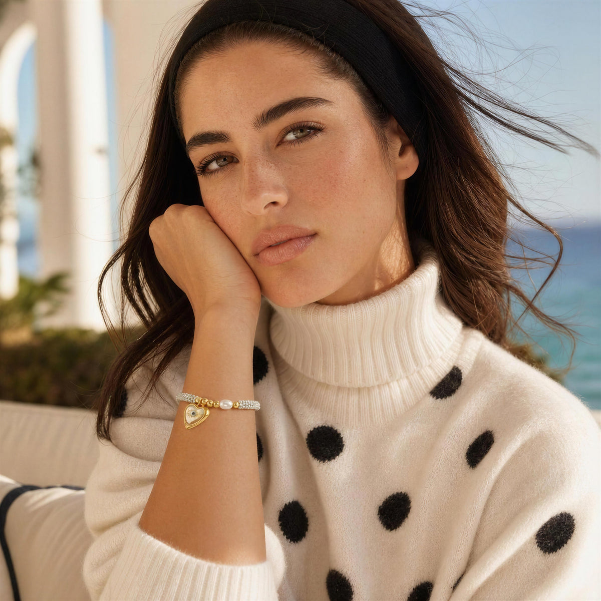 A woman wearing a delicate gold bracelet with a small heart charm and a clear crystal accent, paired with a white turtleneck sweater featuring black polka dots. She is posed outdoors with a blurred background of a beach and white columns.