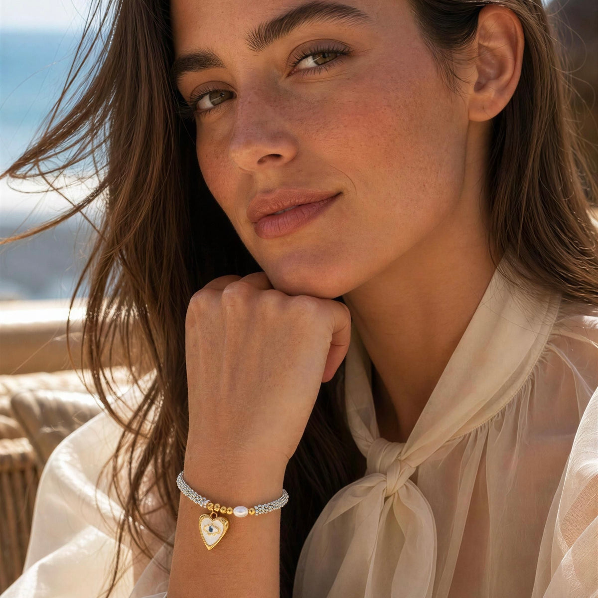 A close-up lifestyle shot of a woman wearing a delicate silver chain bracelet with a gold heart-shaped charm and a white pearl accent, set against a soft-focus beach background.