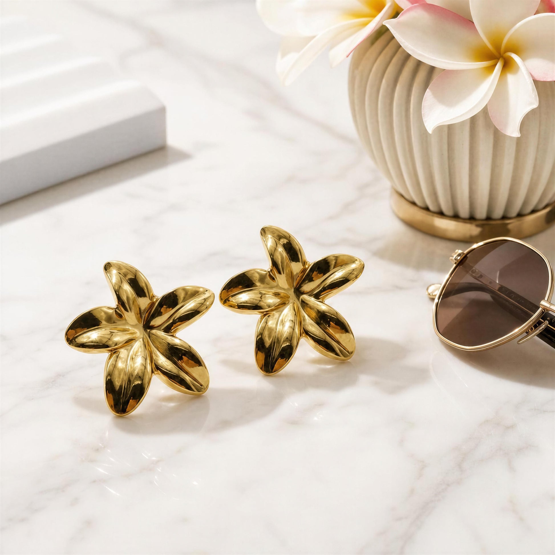 Gold flower-shaped earrings on a marble surface with a vase of flowers in the background.