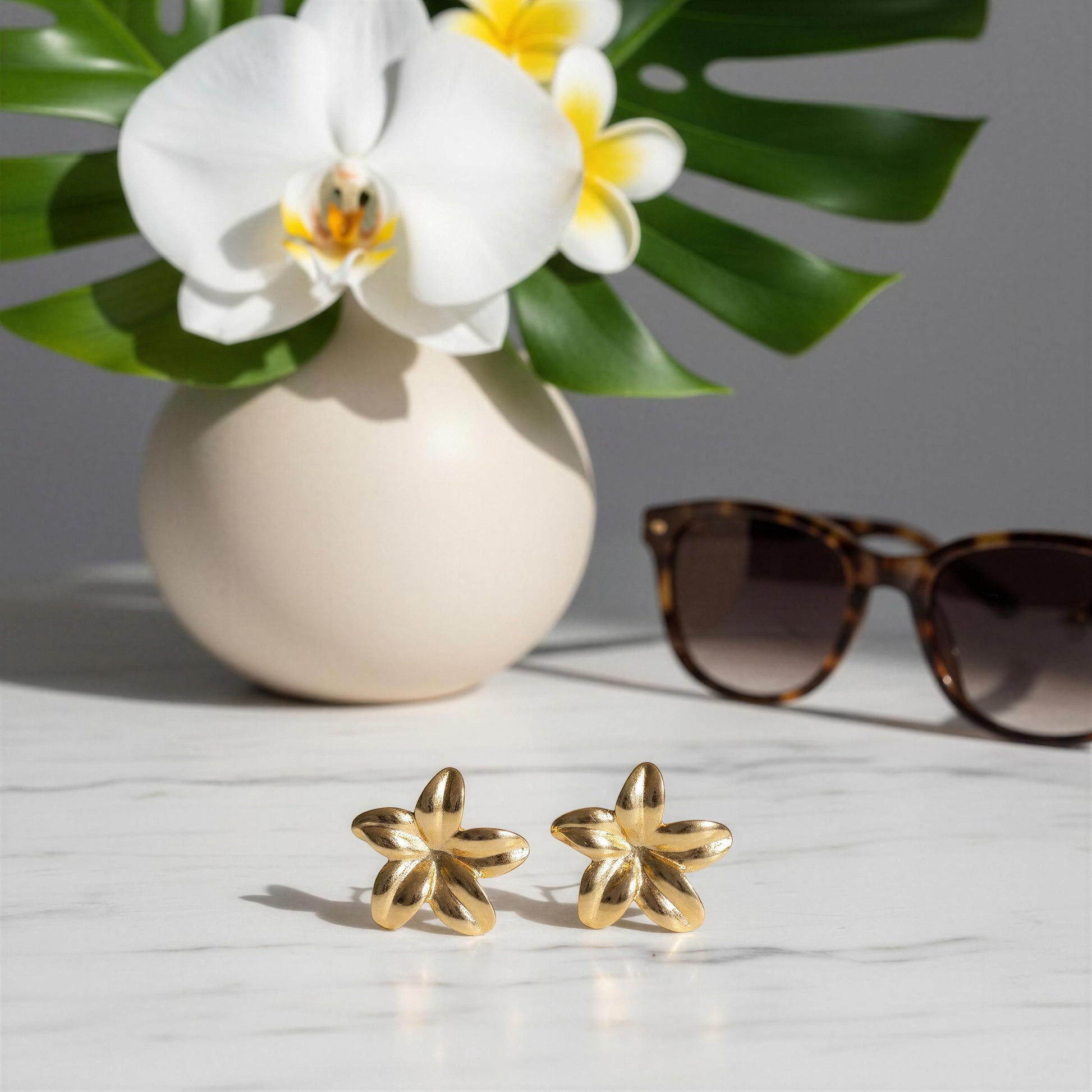 Gold earrings on a marble surface with a vase of flowers and sunglasses in the background.