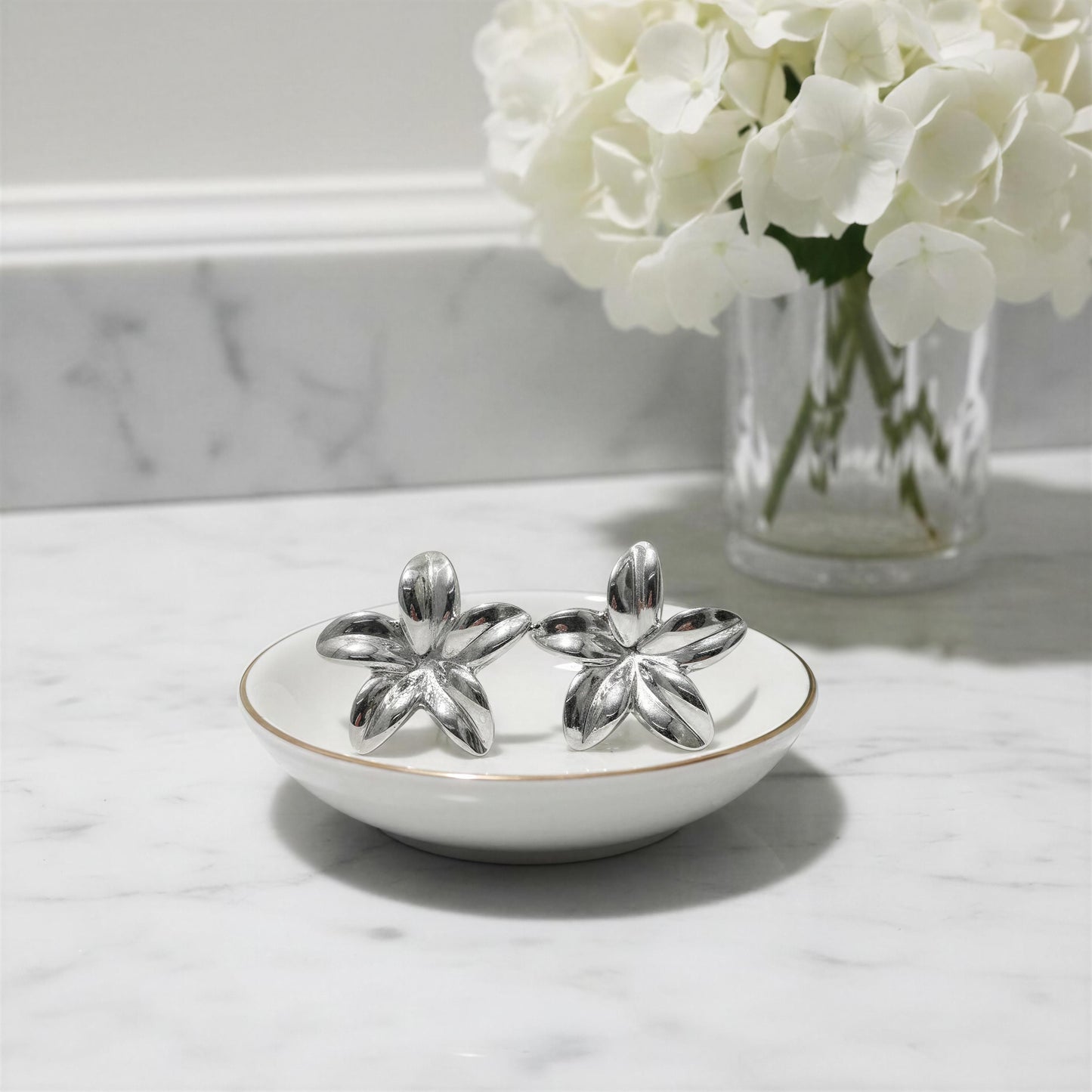 The Luna Bloom Earrings - Silver on a white dish with a vase of white flowers in the background.