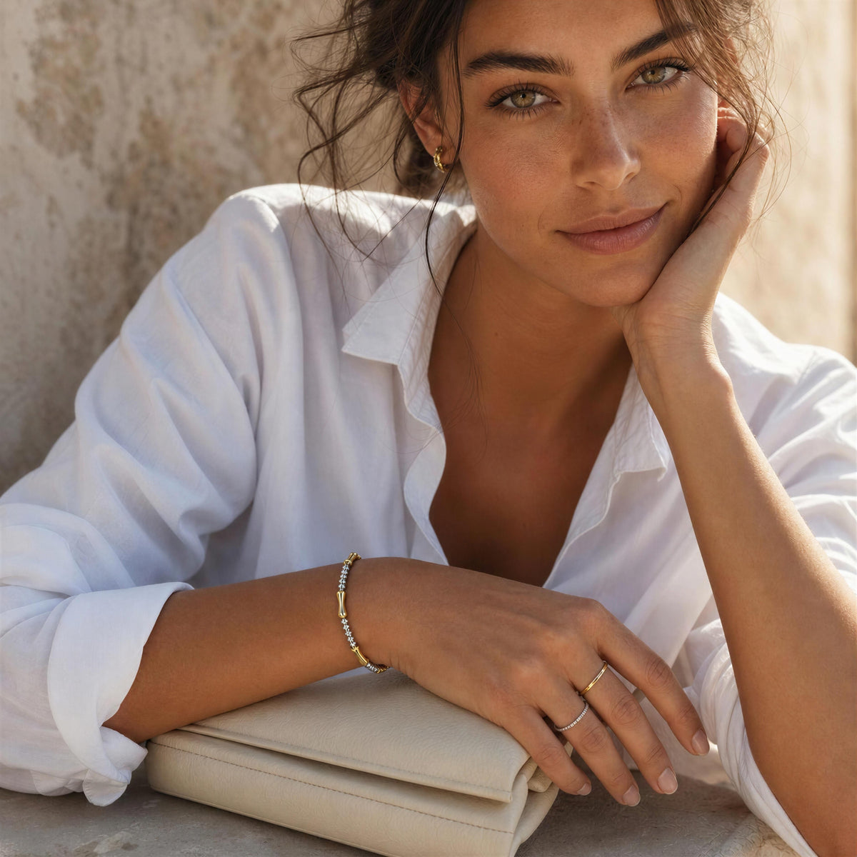 A woman in a white button-down shirt rests her head on her hand, wearing delicate gold rings and the  Malta Bracelet from Chica Chica Jewelry, with a beige clutch bag in the foreground.