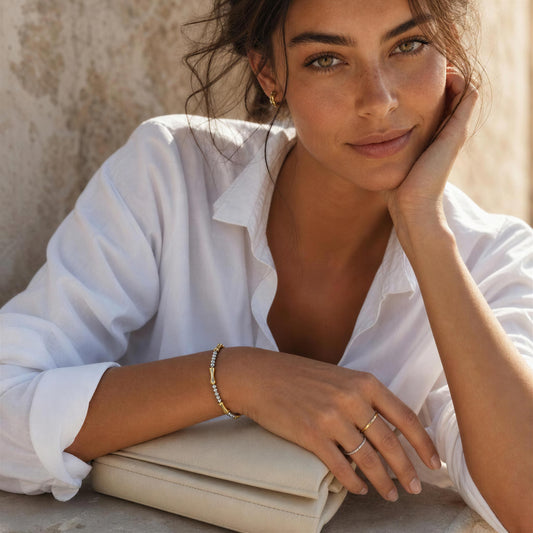 A woman in a white button-down shirt rests her head on her hand, wearing delicate gold rings and the  Malta Bracelet from Chica Chica Jewelry, with a beige clutch bag in the foreground.