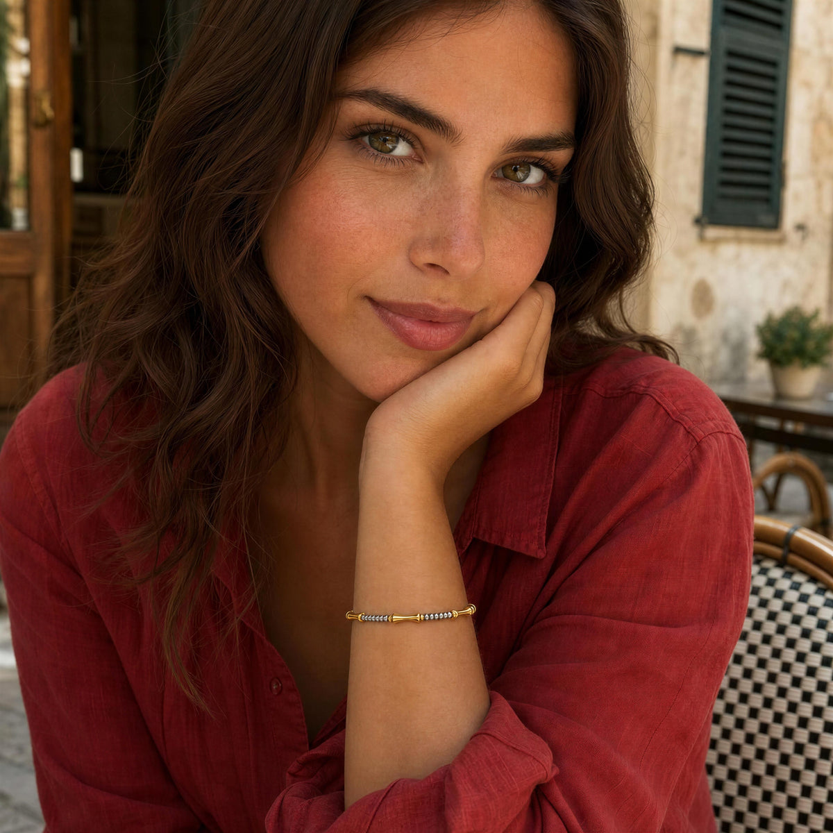 A woman with long brown hair rests her chin on her hand, wearing a delicate gold beaded bracelet on her wrist. She is wearing a red button-up shirt and is seated in an outdoor patio setting with a blurred building background.