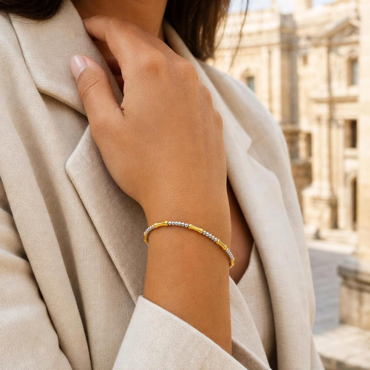 A close-up of a woman wearing a delicate beaded bracelet on her wrist, dressed in a beige blazer with a blurred classical building in the background.