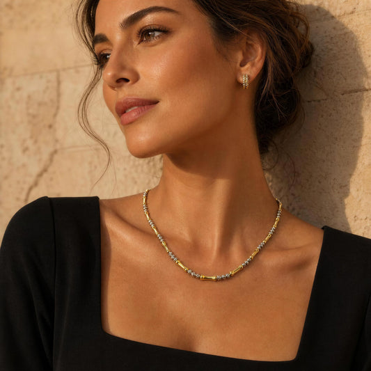 A woman wearing the Malta Necklace from Chica Chica Jewelry , a delicate gold and silver necklace with a matching stud earring, posing against a textured beige stone wall.