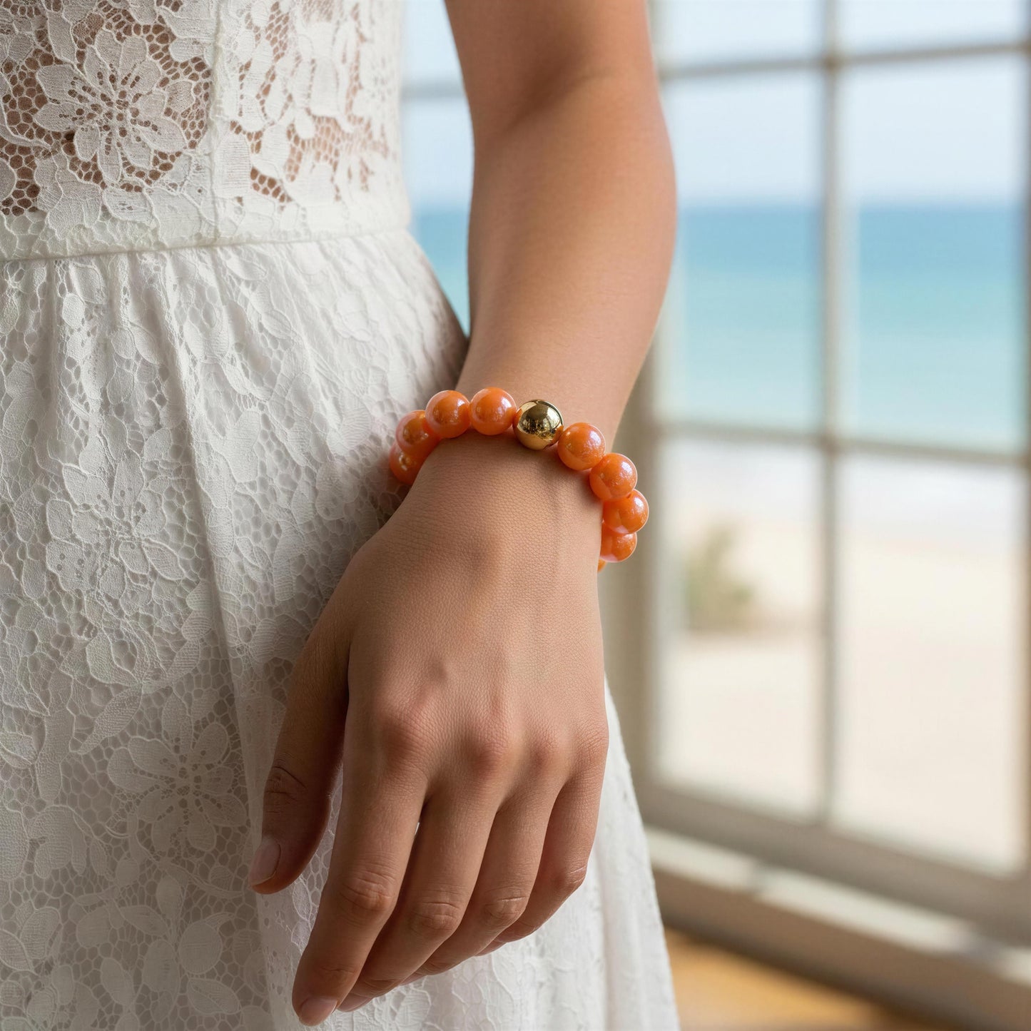 Person wearing an orange bracelet with a gold bead, standing by a window with a blurred outdoor view.