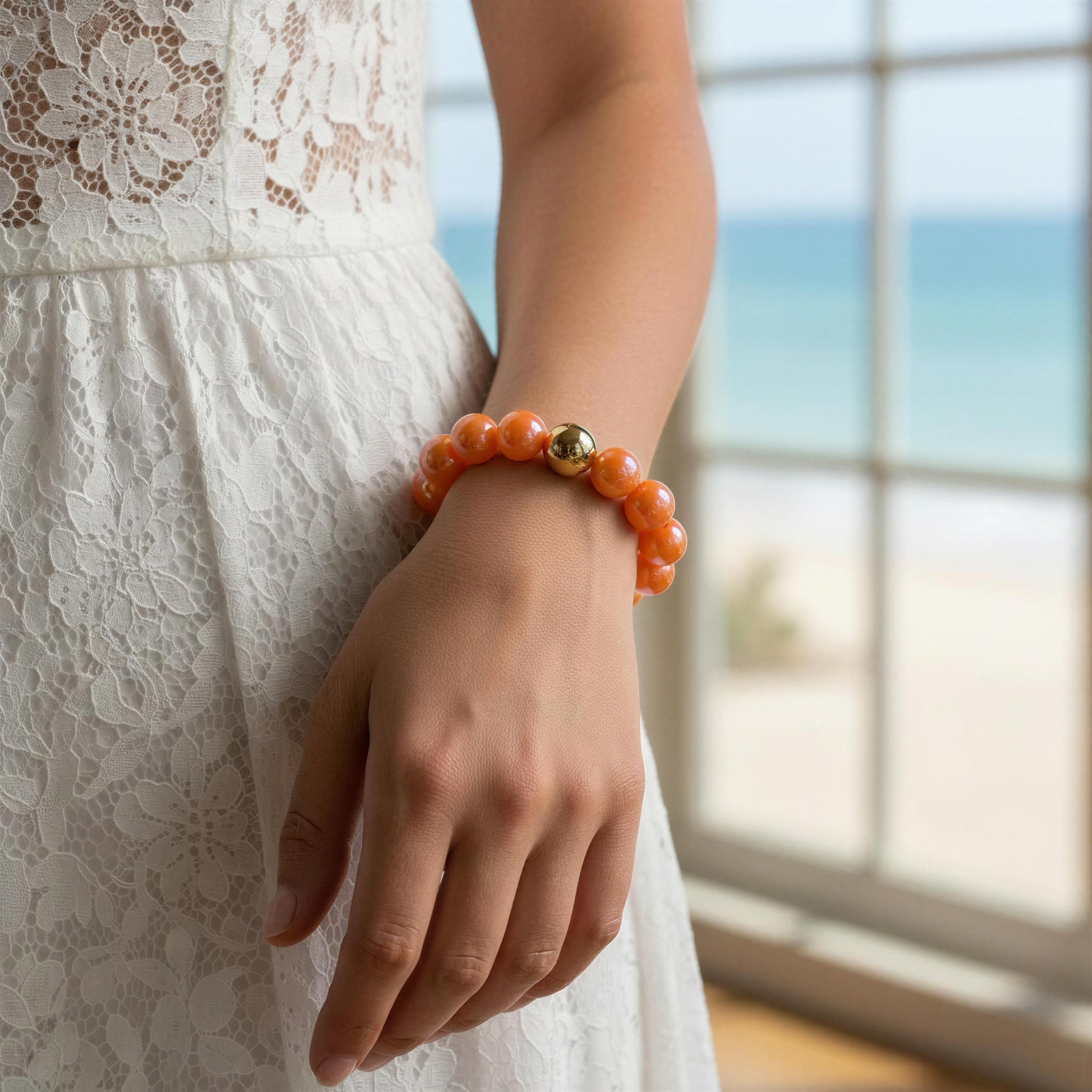 Person wearing an orange bracelet with a gold bead, standing by a window with a blurred outdoor view.