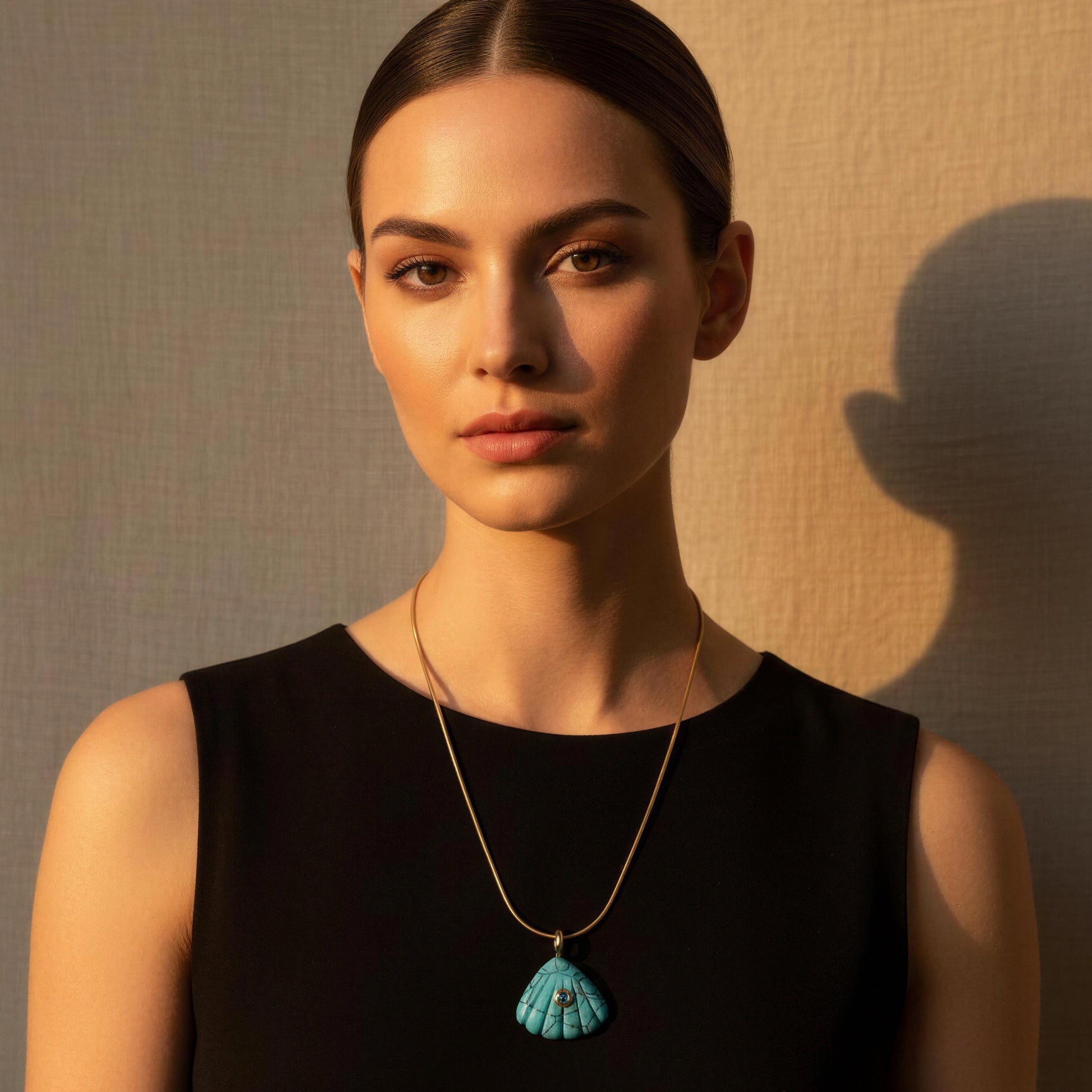 Woman wearing a black dress with a gold necklace and blue shell pendant against a beige wall.