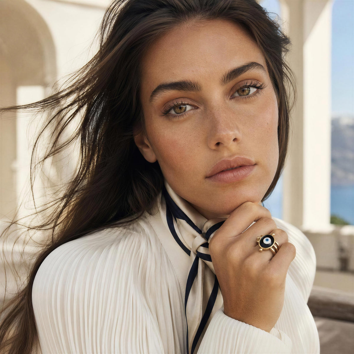 A close-up lifestyle shot of a woman with long dark hair wearing a white pleated blouse and a navy ribbon tie, showcasing a gold ring with a black and white evil eye design on her finger.