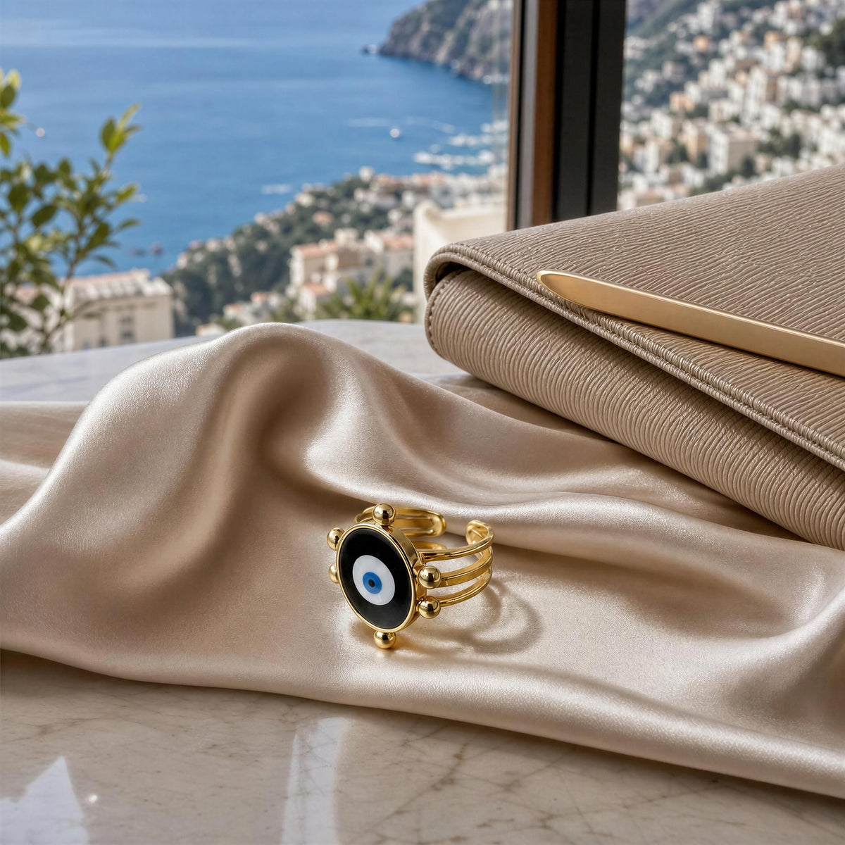 The Mati Ring from Chica Chica Jewelry, styled on draped champagne silk fabric next to a beige clutch bag, with a scenic blue sea and coastal town in the background.
