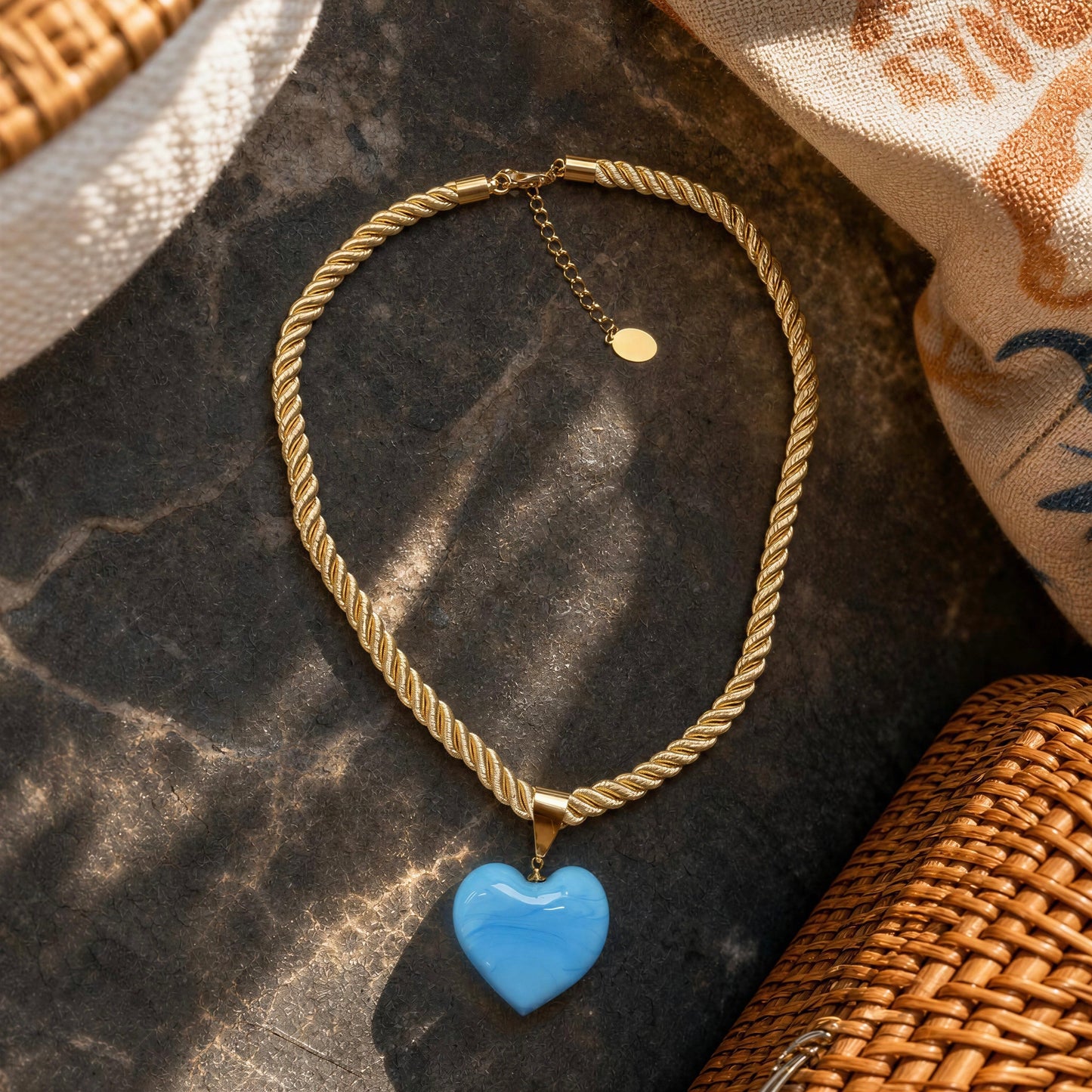 The Mavi Murano-style blue glass heart necklace with a soft braided cord and 18k gold-plated details by Chica Chica Jewelry flat lay on black stone.