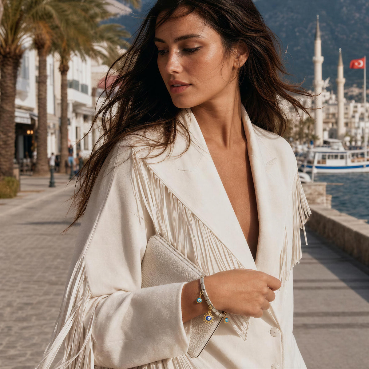 A woman walking along a seaside promenade wearing a cream-colored fringed jacket and holding a matching clutch bag, with a boat and minarets in the background. She wears the Nazar Bracelet
from Chica Chica Jewelry. 