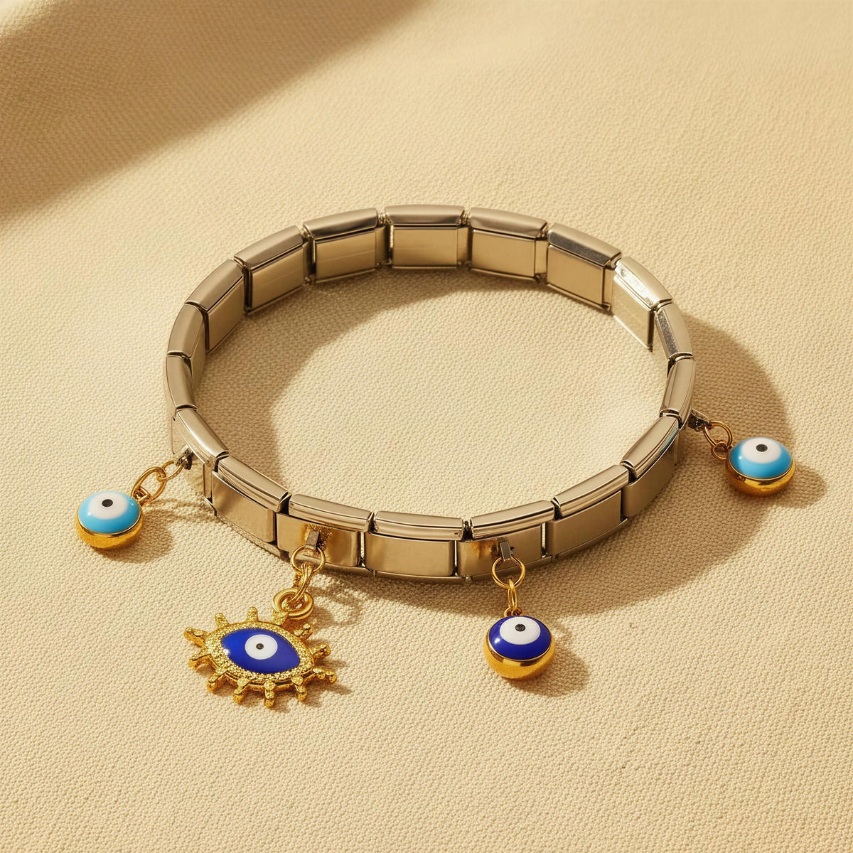 The Nazar Bracelet from Chica Chica Jewelry. A Gold-tone metal link bracelet with three evil eye charms, including one larger sun-and-eye charm, displayed on a textured beige surface.