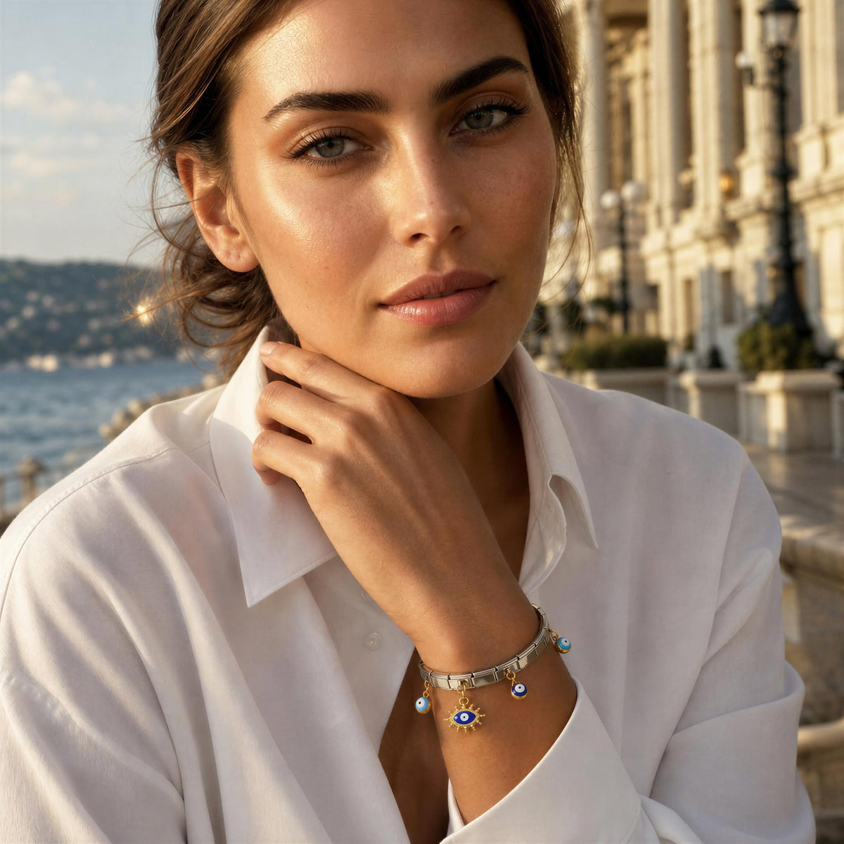 A woman in a white collared shirt wearing the Nazar Bracelet
from Chica Chica Jewelry,   a delicate silver charm bracelet with blue and gold details, posed outdoors against a blurred background of a coastal city and classical architecture.