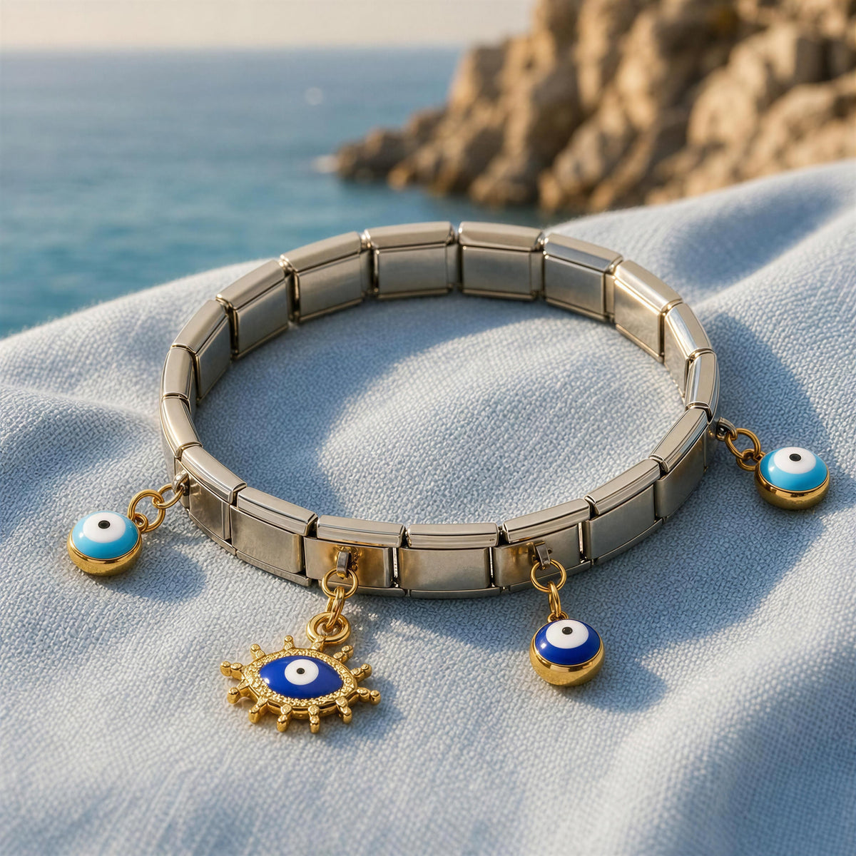 A silver-tone metal link bracelet with three dangling charms, including two blue-and-white evil eye beads and a gold sun-and-eye pendant, displayed on light blue fabric with a blurred coastal background.