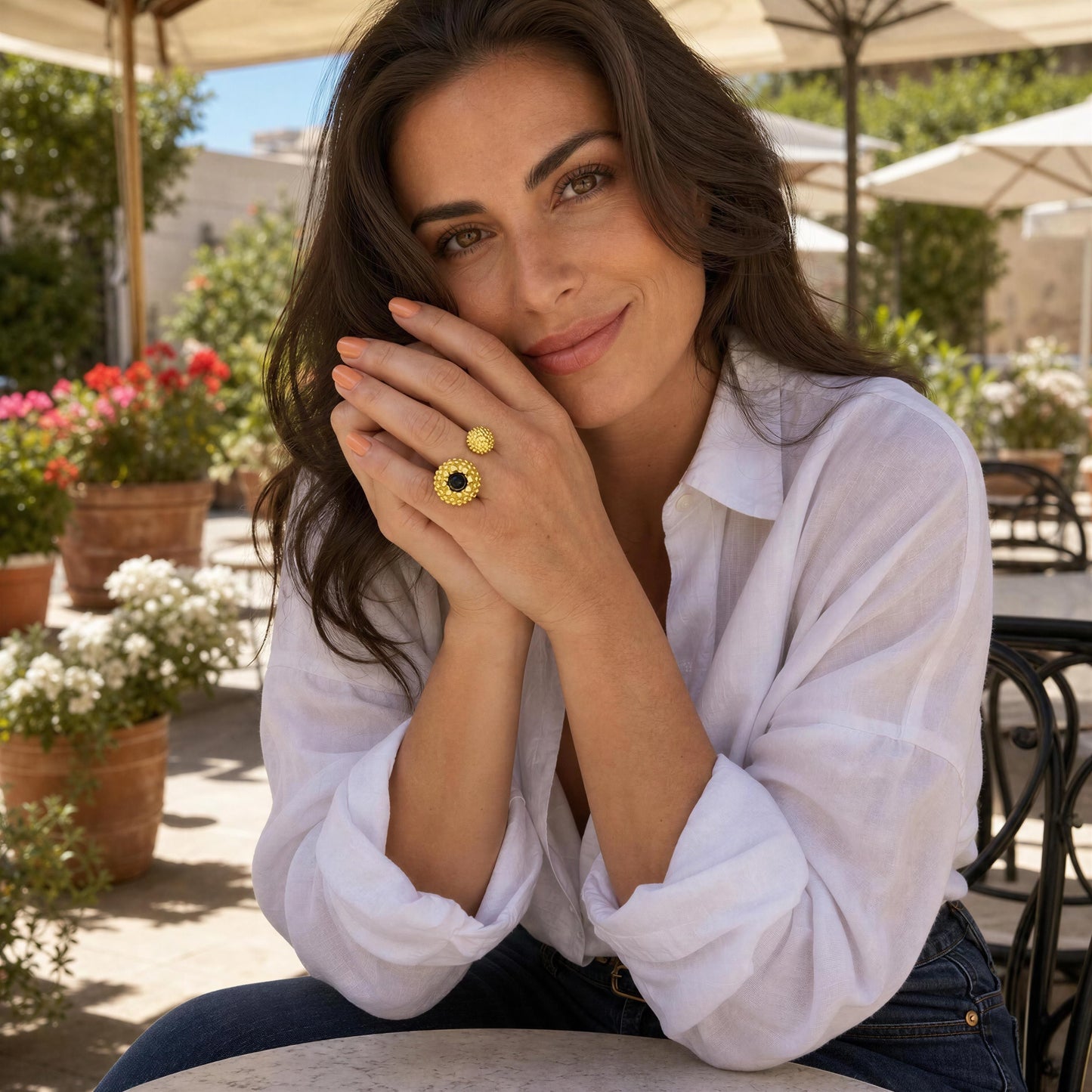 A woman in a white button-down shirt and blue jeans sits outdoors at a cafe table, wearing the Nero Ring from Chica Chica Jewelry. 
