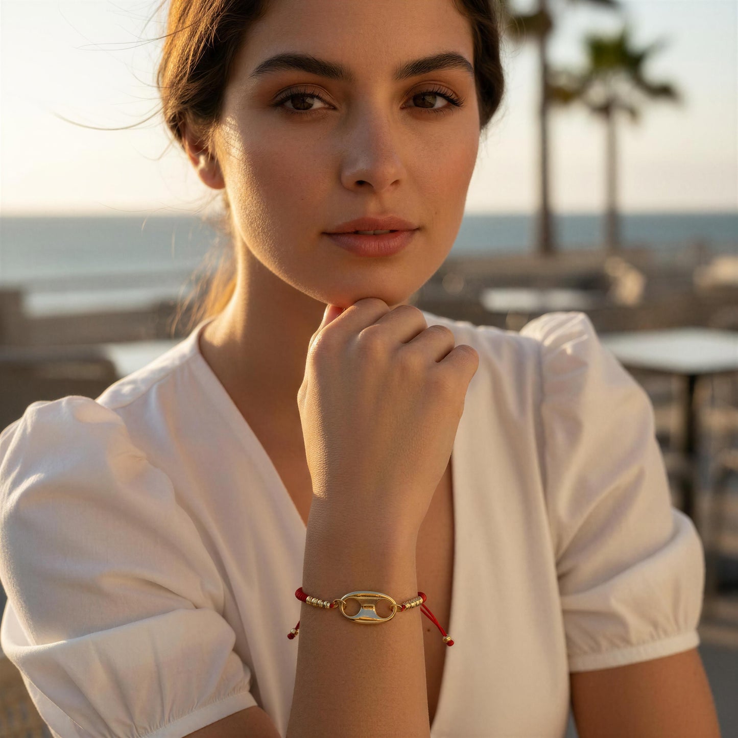 Woman wearing a gold bracelet with a red string, sitting by a waterfront.