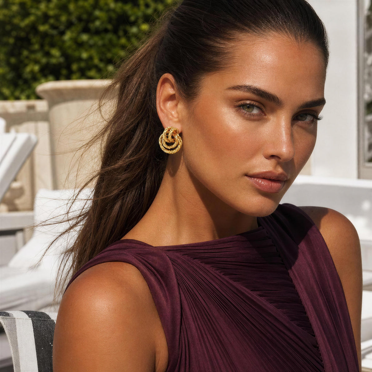 A woman wearing a pair of gold double-helix style hoop earrings, photographed outdoors with a white building and greenery in the background.