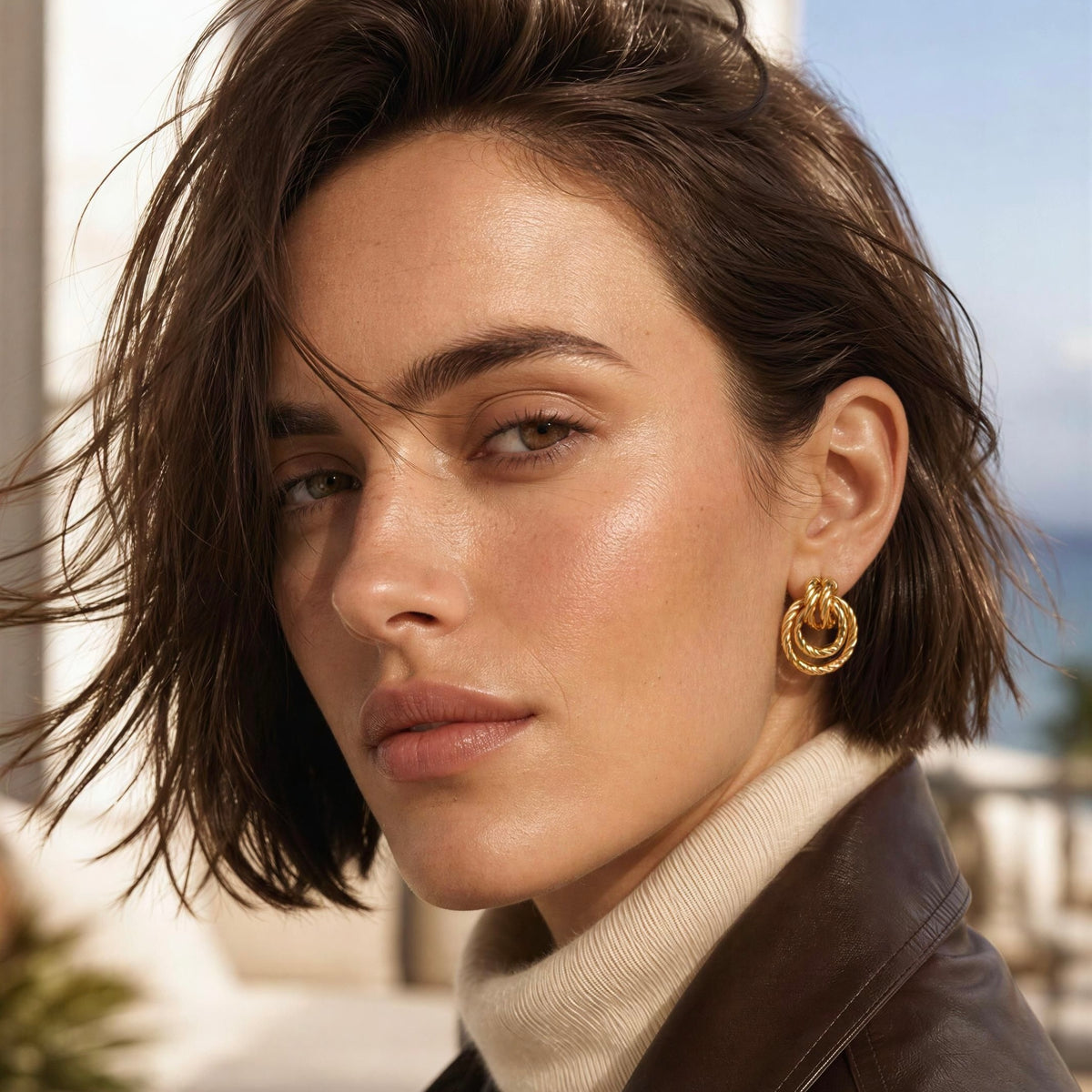 Model wearing the Nodo earrings from Chica Chica Jewelry and a leather chocolate brown jacket and creamy cashmere turtleneck by outdoor terrace. 
