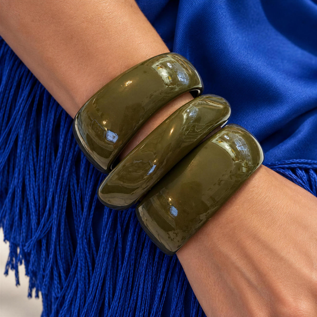 The Oliva Bangle Set from Chica Chica Jewelry. Three  glossy olive green bangle bracelets stacked on a person's wrist, with a bright blue fringed garment in the background.