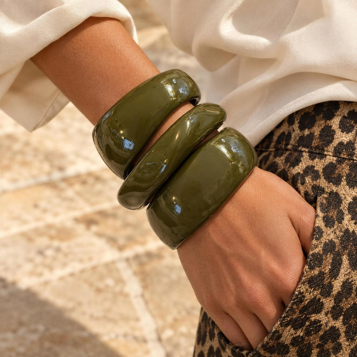 The Oliva Bangle Set from Chica Chica Jewelry.  Three glossy olive green ceramic bangles stacked on a person's wrist, shown with a white blouse and leopard-print pants against a sunlit stone background.
