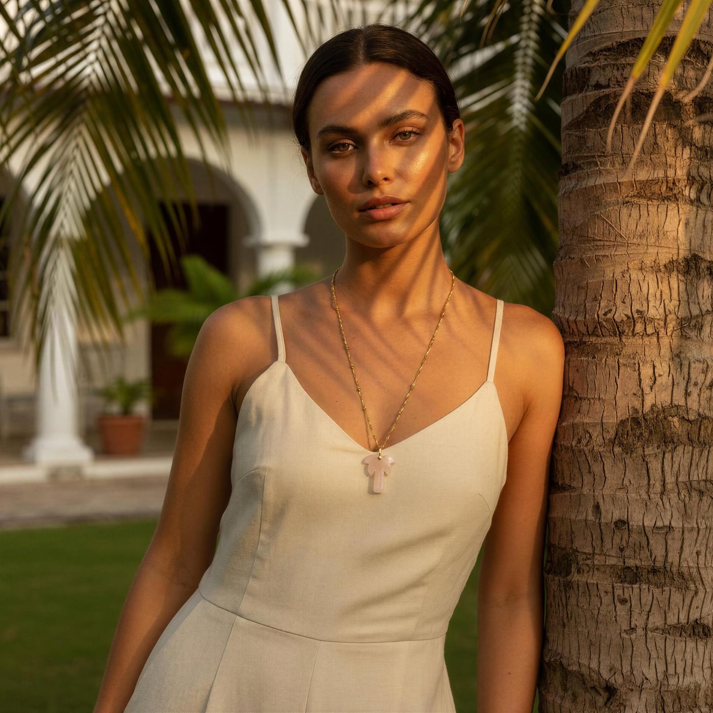 Woman in a beige dress standing next to a palm tree with a warm, natural light wearing the Palma Rosa Long Necklace