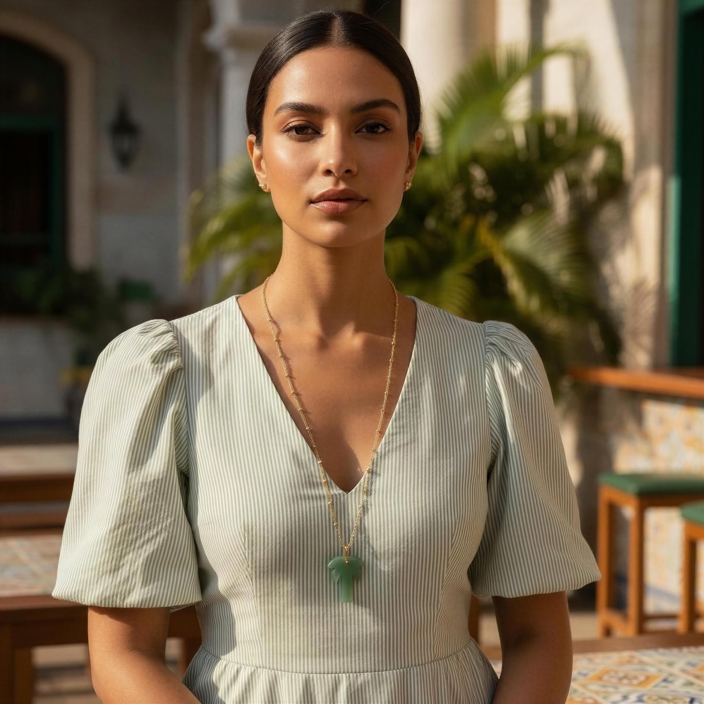 Woman in a light green dress standing outdoors with plants and a building in the background wearing the Palma Verde Long Necklace