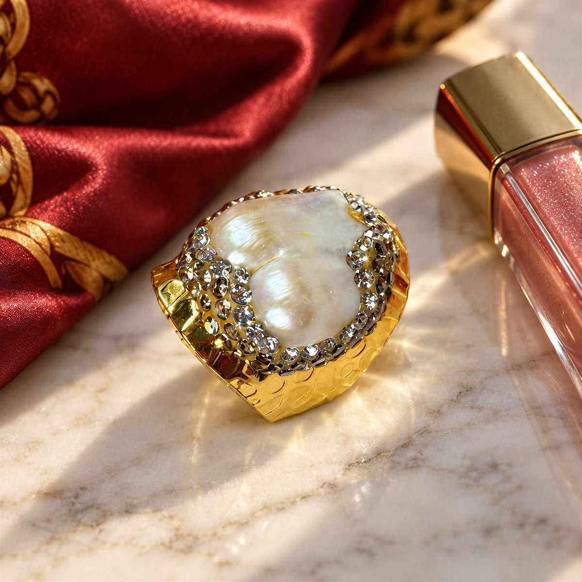 The Perla Ring from Chica Chica Jewelry, a gold ring featuring a large white mother-of-pearl cabochon and a border of clear rhinestones, styled on a marble surface next to a red satin fabric and a pink lip gloss bottle.