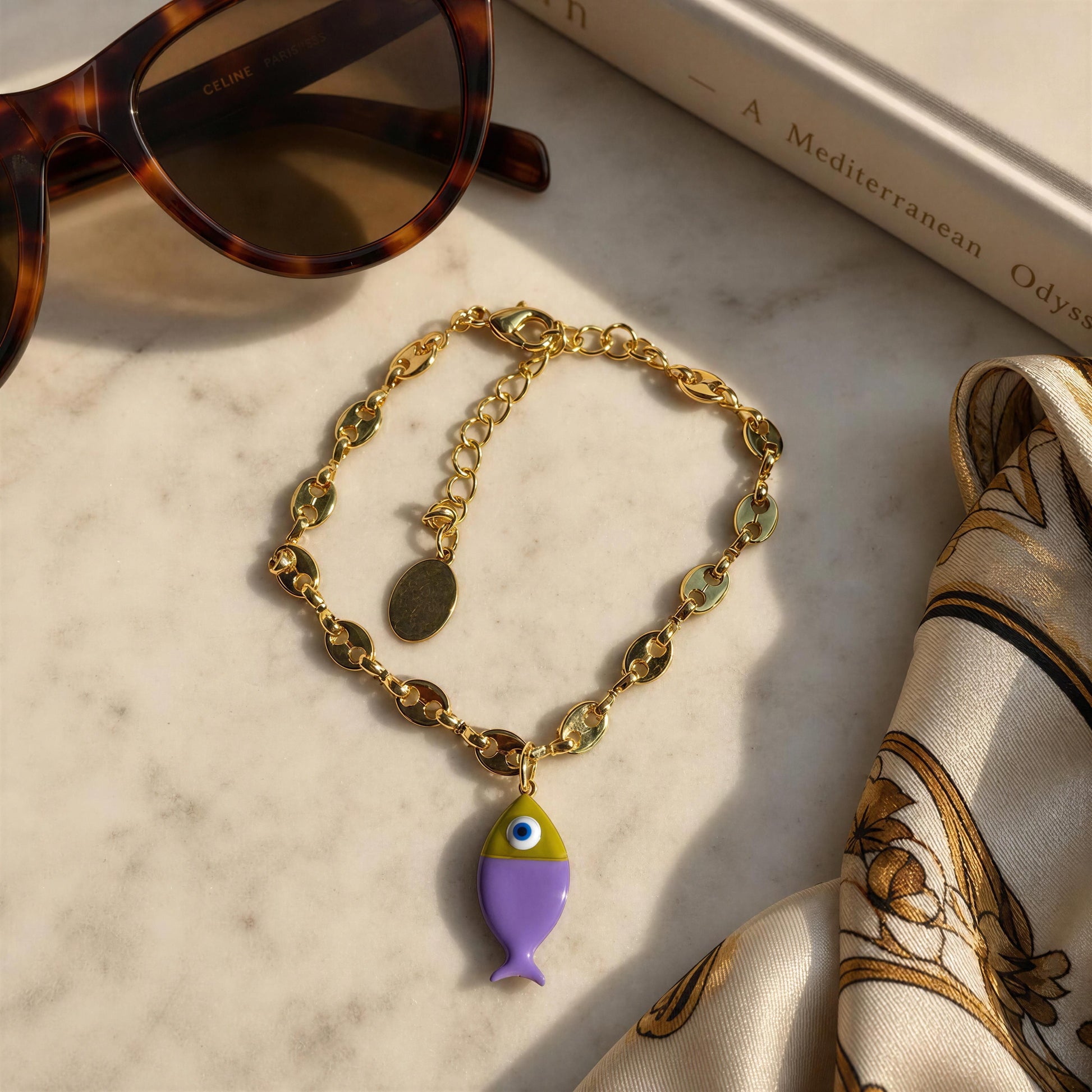 The Pesca Lila Bracelet with a fish charm on a marble surface with sunglasses and a book.